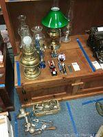 Seven hurricane lamps with glass chimneys and green metal shades shown on wooden furniture surface, accompanied by small items and three bar tap handles.