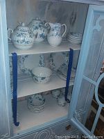 Porcelain china pieces arranged in a white cabinet, showing variety of plates, bowls, tea cups and serving dishes all with blue floral pattern.