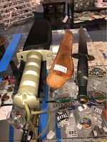 Photo of both knives and their sheaths on a glass display. The fighting knife shows a metallic striped handle and a black sheath. The survival knife displays rust and a brown leather sheath.