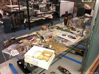 Wide view of glass display shelf showing assorted vintage collectibles including toy guns, figurines, knives, and paper ephemera arranged under glass.