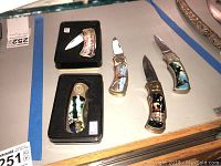 View of five folding knives with decorative western and nature-themed handles, three out of five shown with blades open, two knives in display tins.