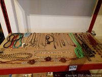 Top-down view of a collection of various costume jewelry necklaces laid out on a table, showing multiple styles, colors, and pendant designs including a prominent white cross pendant on a blue beaded necklace.