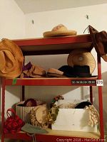 Full view of red metal shelf displaying a collection of vintage hats including straw and felt hats, and hat accessories, arranged over two shelves and the box below.
