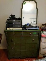 Full front view of vintage green chest of drawers with mirror showing the drawer configuration and top surface with some items set on it.