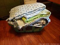 Stack of bedding items in a green laundry basket, showing folded bedspreads and blankets in coordinated colors and patterns.