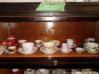 Full display of multiple tea cups and saucers on a wooden shelf, showing varied designs and colors including red, pink, and gold accents.