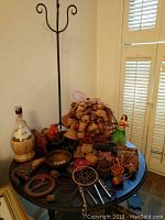 Display of numerous small woven baskets, decorative miniature hats, a hula girl figurine, glass bottle with wicker cover, and other small ornamental items on a round wooden table beside a tall, black metal stand.