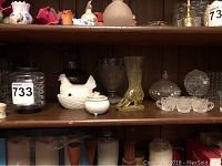 Shelving with various glassware items including a yellow glass boot vase, cut glass candy dishes shaped like hens with lids, and other decorative glass pieces including children's tea cups and votives.