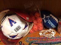 Photo showing red leather soccer ball holder with white/red soccer ball, plastic bag of orange wiffle balls, blue Dallas Cowboys football, strand of shell necklace and colorful bead necklaces on wooden shelf.