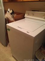Front and side view of a white top load washing machine in a laundry area, showing the lid closed and machine body.