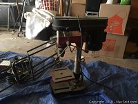 Full side view of the 5 speed drill press placed on tarp showing metal base, work table, control arm, and power cord.