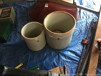 Two vintage stoneware crocks placed on a blue tarp with surrounding tools and items, showing overall size comparison and condition.