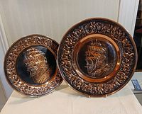 Pair of copper ship wall plaques displayed side by side on a white surface, showing size difference and details of design.