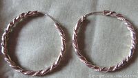 Pair of twisted sterling silver pierced hoop earrings on a beige fabric background, showing round hoops with detailed twisted pattern and secure clasp.