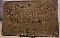 Front view of brass shipbuilder plaque with embossed lettering and decorative corner elements.