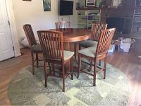 Full view of the wood round high-top table with five wooden chairs arranged around it, placed on the green patterned round rug.