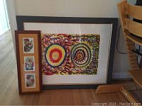 Framed signed original painting with abstract circular designs and framed triptych floral print leaning against the wall on wooden floor near chair.