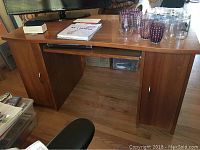Front view of wood desk with pull-out keyboard tray under the desktop, two side compartments each with a door, large flat surface with glassware and papers on top.