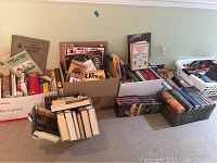 Wide view of several boxes and baskets of assorted books and boxed sets on the floor showing variety of titles and subjects.