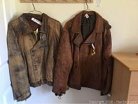 Two leather jackets hanging on hangers side by side against a wall, showing full front and some side view.