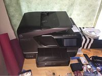 Photo showing the HP Officejet Pro 8600 Plus wireless printer on a wooden surface with surrounding small items and a green curtain in background.