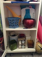 Shelf with blue ceramic pot with tray, orange watering can, red ceramic pot, small plastic vase, and four small glass containers.