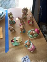 Overview of nine hand painted Pendelfin rabbit figurines on wooden table, various poses and outfits with soft pastel colors.