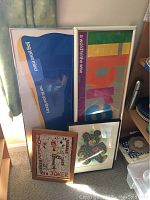 Four pieces of art leaning against a wall near shelves, various sizes and colors, two large text-based prints, one small bear print, one medium signed Joker mixed media.