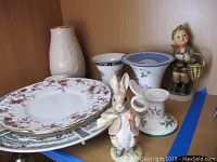 Porcelain and ceramic items arranged on a wooden shelf showing Lenox vase, Wedgewood plates and small vases, Goebel rabbit figurine, and Royal Albert figurine