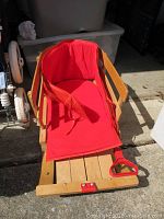 Wooden baby sled with red vinyl padded seat and backrest, safety strap, wooden side rails, red plastic front handle, positioned indoors on concrete floor.