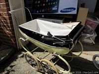Black and green painted metal vintage doll stroller with a white interior and detached shade inside, shown from a side angle outdoors, displaying the four large spoked wheels with cream-colored tires.