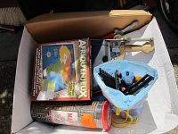 Overview of box containing Afriquenjeux board game, magnetic dart game in container, blue pouch with markers, and two plastic toy swords with foam handles.