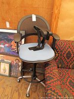 Office chair with tall mesh back, light blue cushioned seat, and spare black plastic armrests on seat, next to a patterned upholstered chair and framed art on floor.