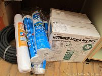 Photo shows two boxes of Gourmet Liquid Ant Bait (Honeydew formula), three rolls of plastic sheeting (two are blue embossed, one frost guard fleece), and black rubber garden hose coiled on floor.