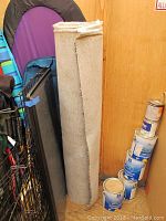 Three rolled segments of Berber carpet leaning against a wall beside paint cans and a folded fabric tent.