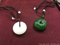 Photo of a pair of jade pendants on black braided cords with bead accents placed on a red surface.