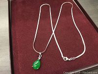 Full view of the green jade teardrop pendant necklace laying on a reddish-brown velvet display surface.