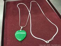 Full view of the green jade heart pendant on a silver snake chain necklace displayed on maroon velvet.