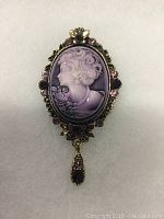 Close-up photo of the purple cameo brooch showing the front detailed relief and surrounding rhinestones.