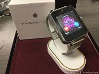 Smart watch on display stand in front of white box labeled Smart Wearable Device. Screen shows time, date, and icons for messaging and contacts.