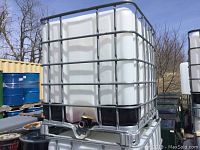 Photo of a large white plastic IBC tote with gray steel cage, mounted on a metal pallet with dispensing valve at the bottom front.