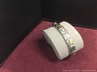 Full view of the silver-tone bracelet displayed on a gray cushion inside a maroon-lined black box. Shows various colorful enamel links.