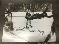 Black and white 16 x 20 print photo of Bobby Orr mid-air scoring a goal with NHL players and goalie visible, including printed facsimile autograph in blue ink.