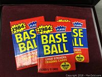 Three unopened 1984 Fleer Baseball Cards packs, red and blue with yellow text showing '1984 Fleer Baseball' and information about logo stickers and photos on each card.