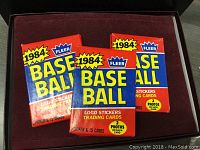 Three sealed packs of 1984 Fleer Baseball cards with bright red and blue packaging featuring logo stickers and trading cards label. Packs are presented on a dark red lined surface.