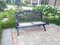 Front view of black wrought iron outdoor bench with wood slat seat on paved patio with greenery background