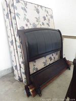 Wooden sleigh bed parts including headboard with leatherette inserts, footboard, wooden side rails, and two floral mattresses stacked against wall.