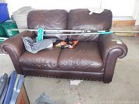 Front view of brown leather loveseat showing two seat and back cushions, rolled arms, tack stud trim, and wear marks including scratches and creases.