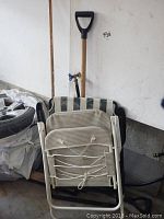 Two folded outdoor folding chairs with metal frames and mesh seating stacked, leaning against a wall with a snow shovel behind.