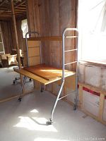 Wood and metal rolling desk positioned near unfinished wall with insulation and wooden framing, showing worn wooden surface and metal frame with wheels.